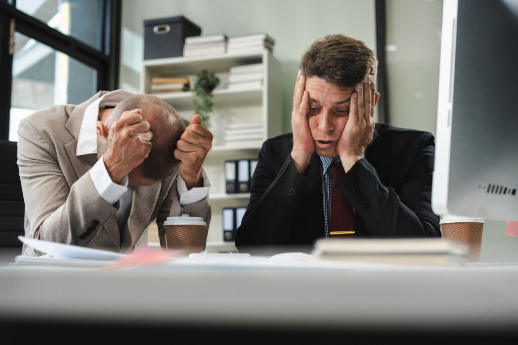 Two stressed businessmen sitting at a desk in an office, looking overwhelmed as they review documents. One man has his head down with clenched fists, while the other holds his face in his hands. The image conveys financial frustration and the challenges of utilizing a poor bookkeeper, reinforcing the need for professional financial management.